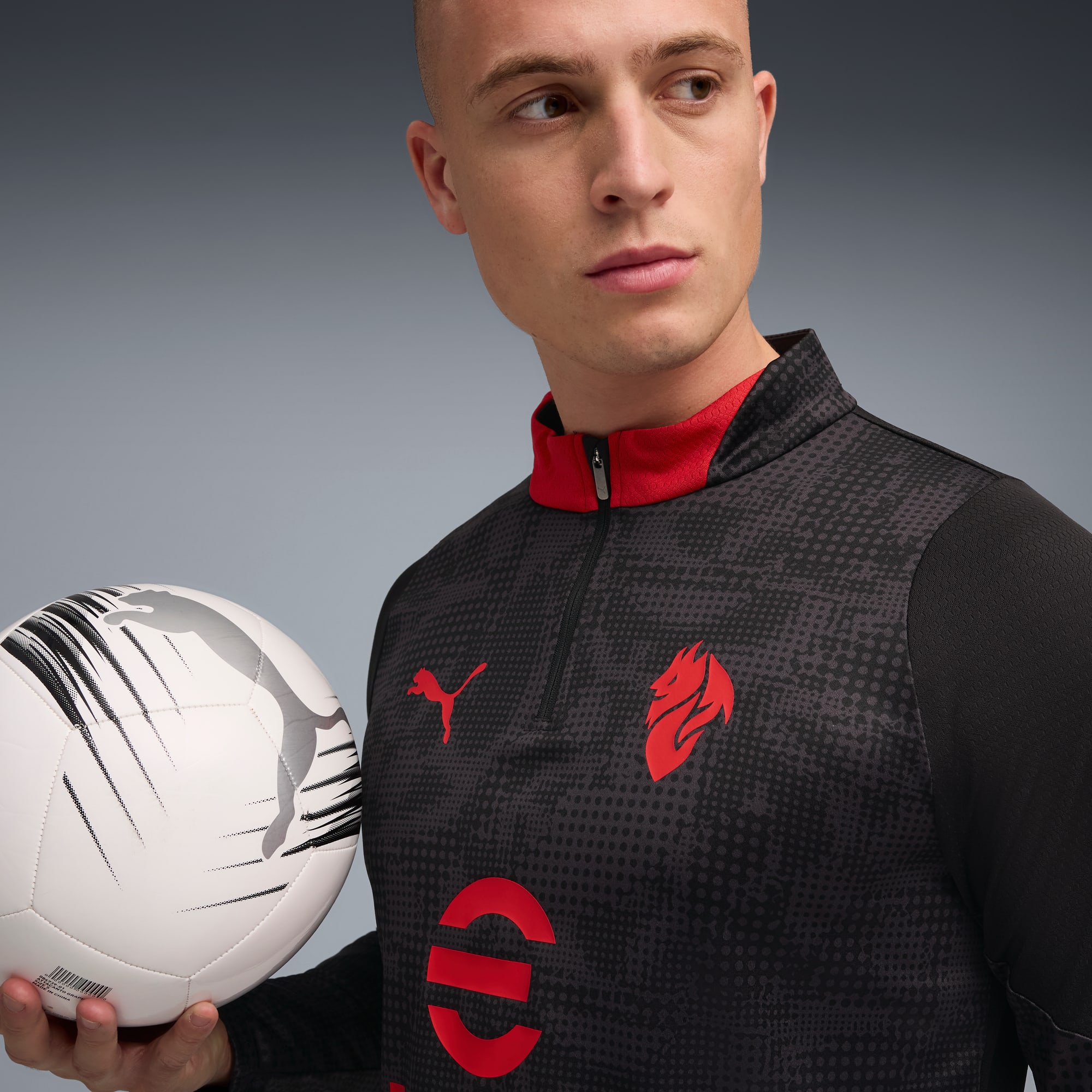 Su sfondo grigio, un modello tiene in mano un pallone e indossa una maglia a motivi neri e rossi.