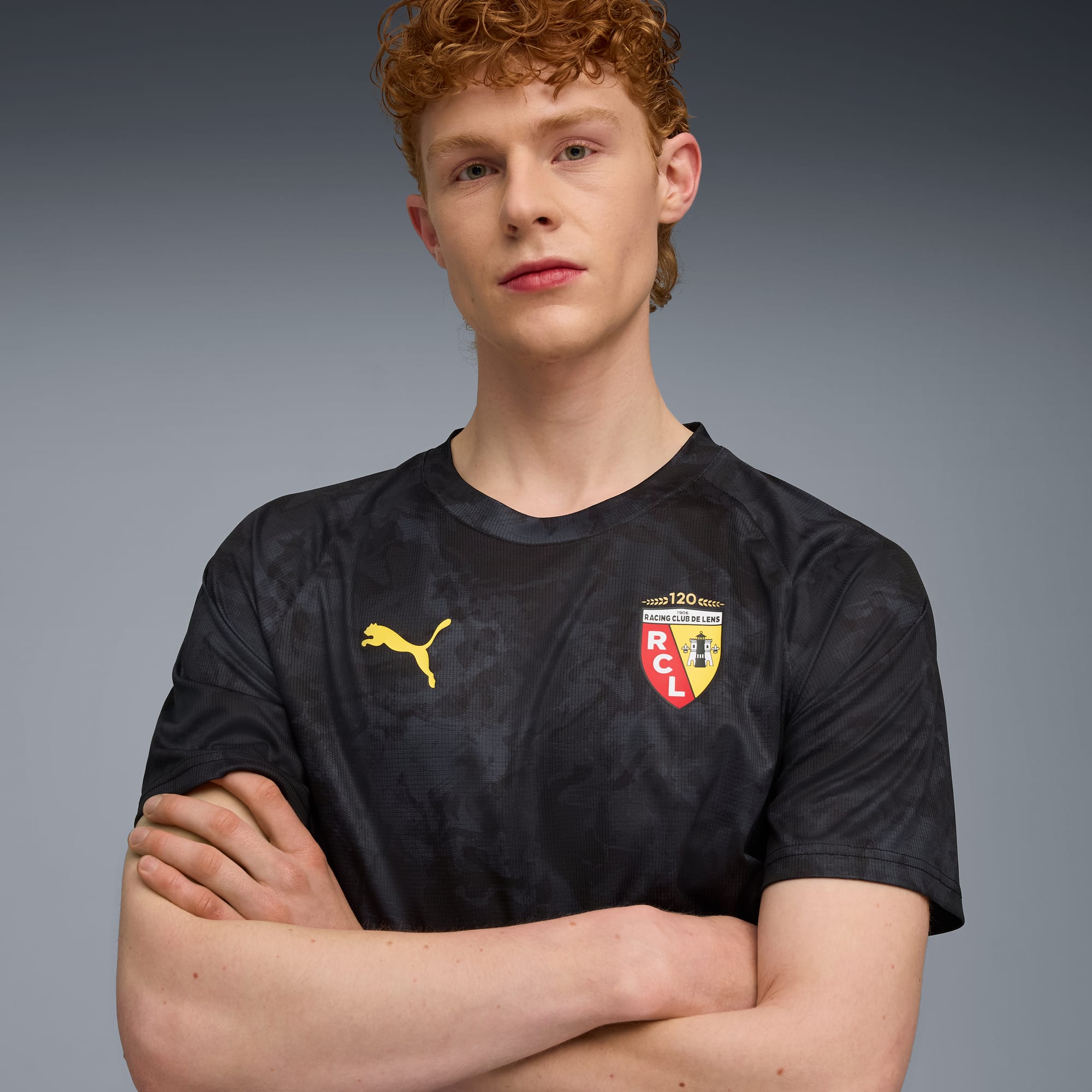 Su sfondo grigio, un modello con le braccia conserte indossa una t-shirt nera con loghi Puma e club.