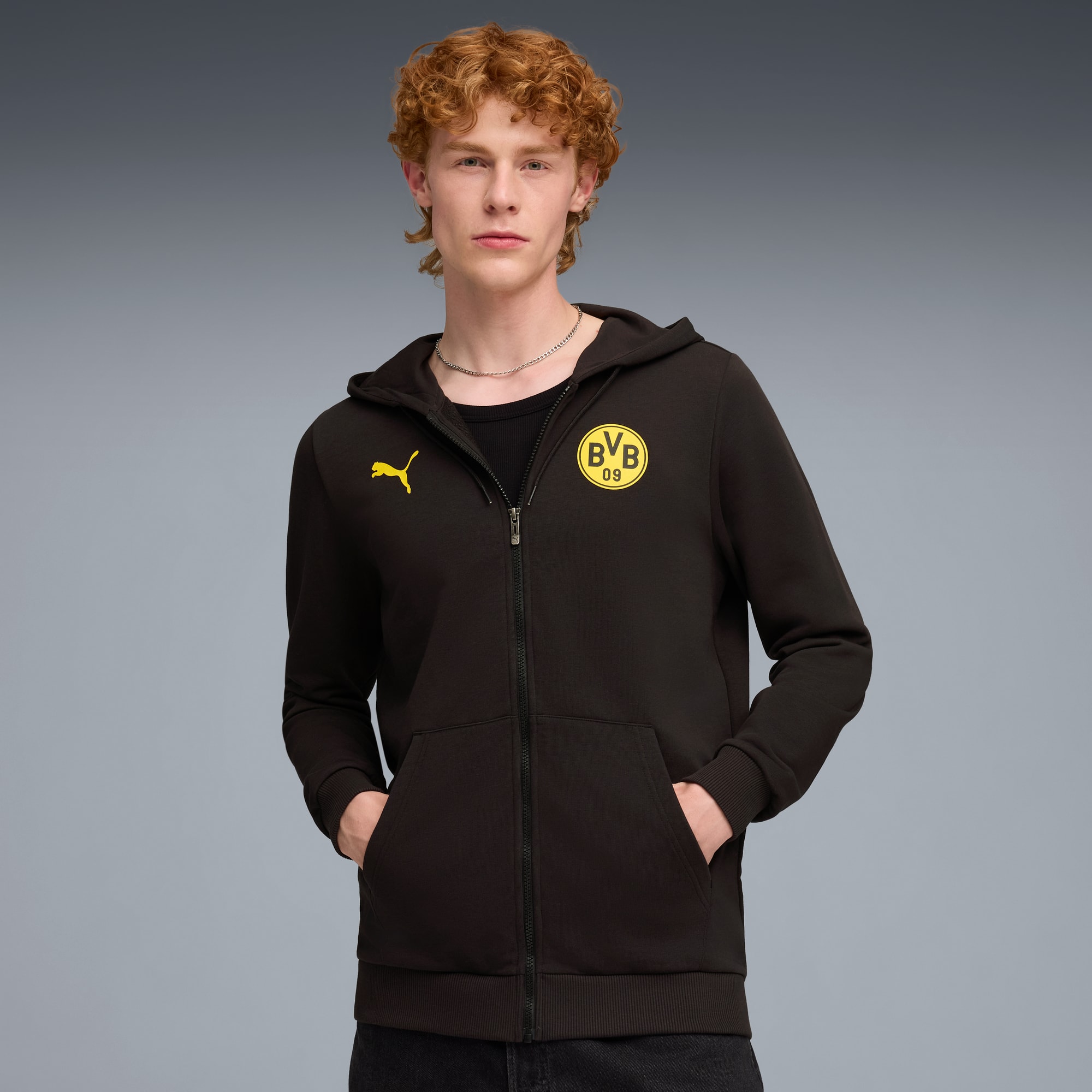 Model pozuje na szarym tle w czarnej bluzie z żółtym logo i dżinsach.