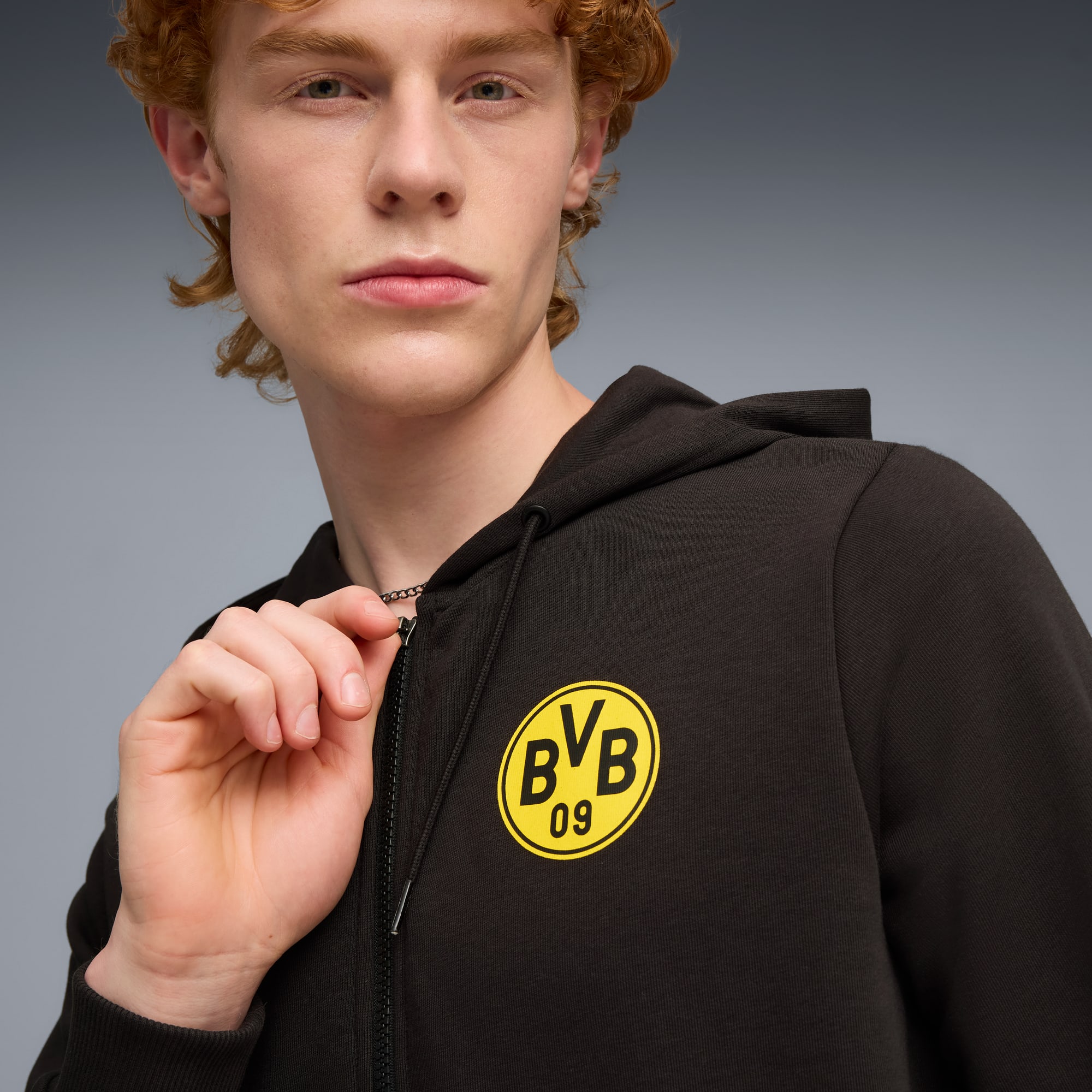 Na szarym tle model poprawia zamek ciemnej bluzy z kapturem z żółtym emblematem.