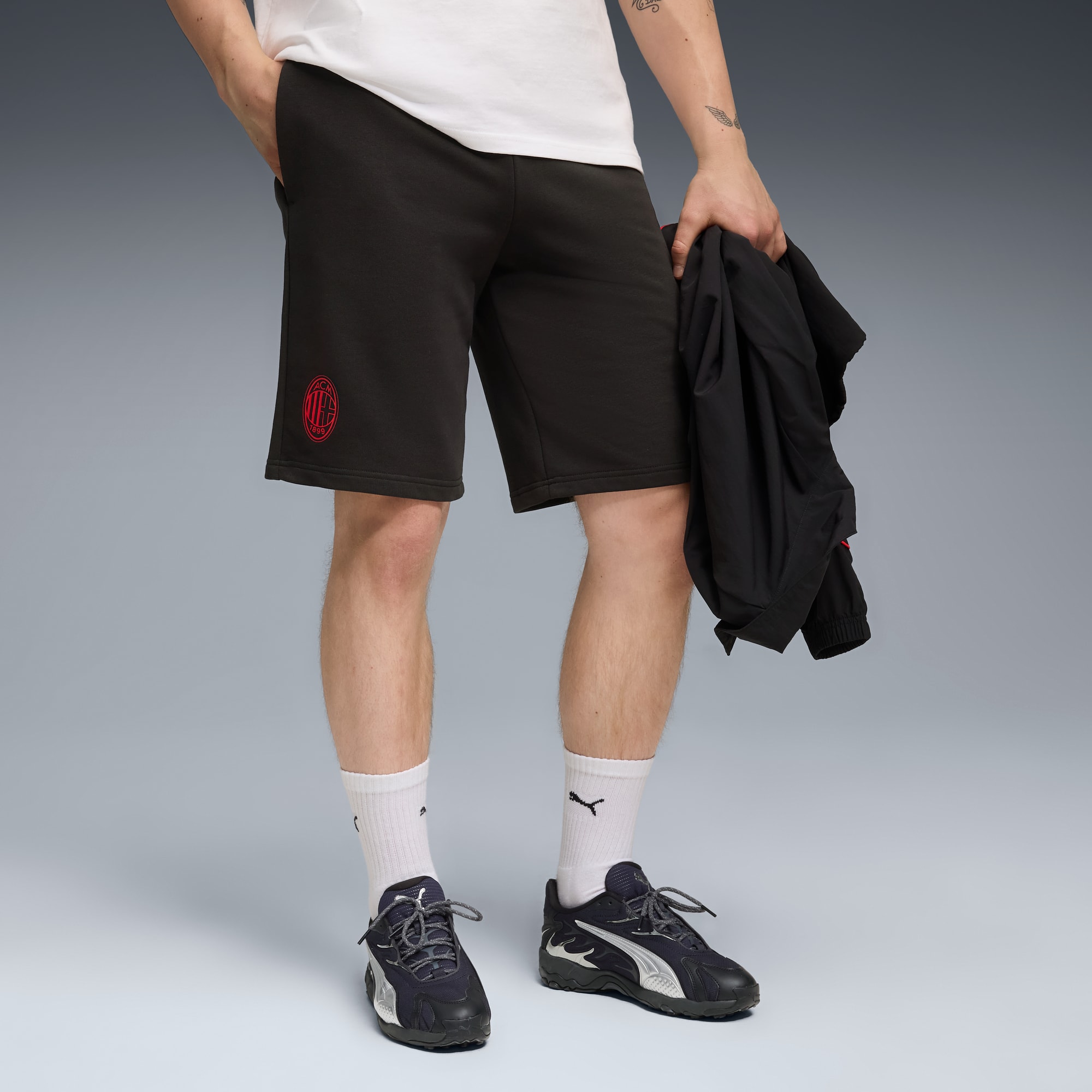 Against a grey backdrop, a model dons black shorts, socks, and trainers, holding a jacket.