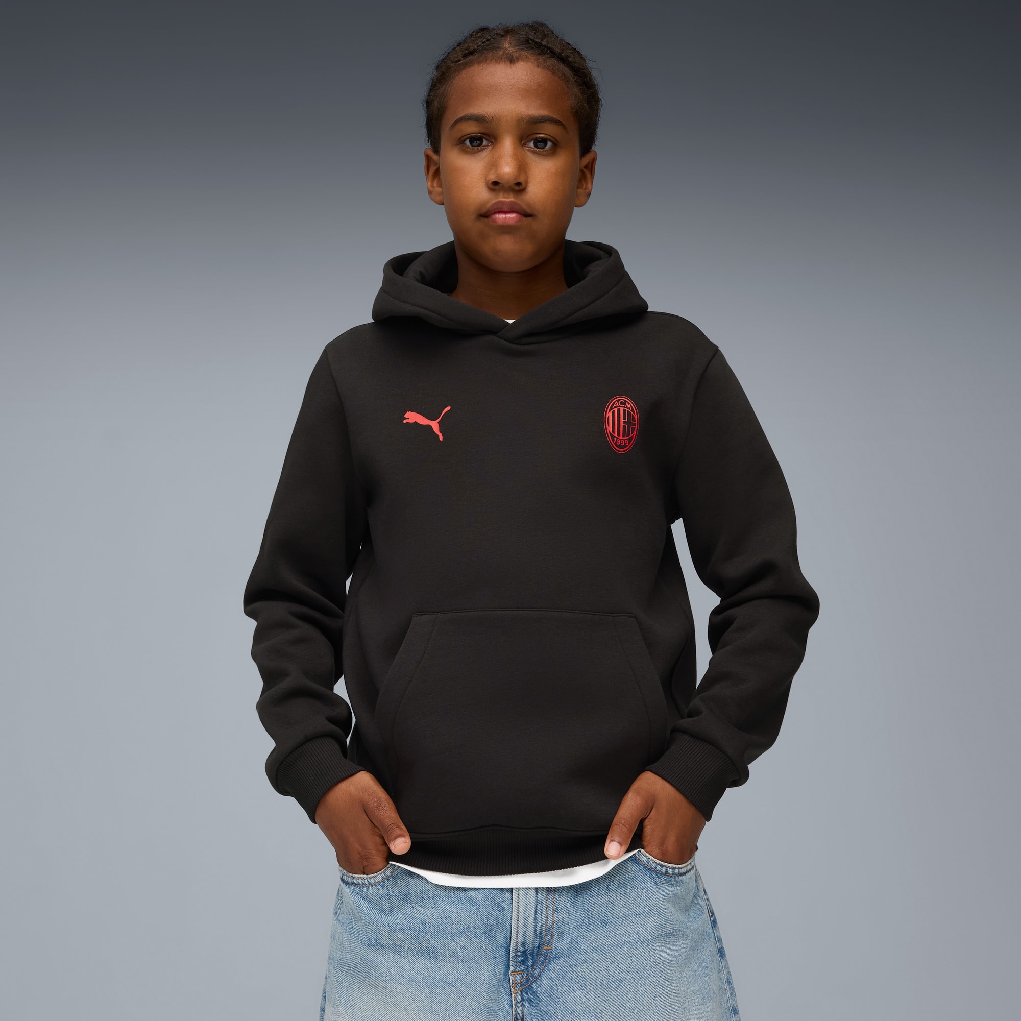 Against grey, a model poses wearing a black hoodie, with red logos and blue jeans.