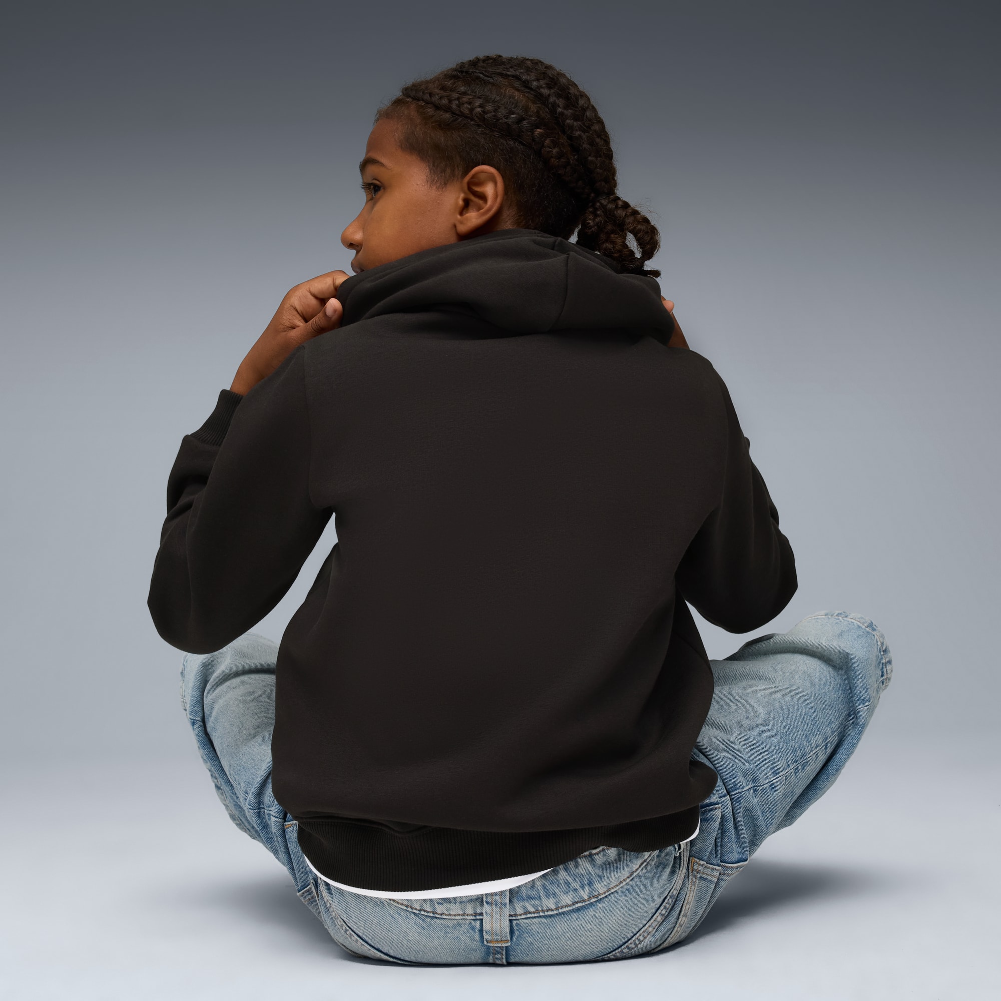 A model with braids, seated against a grey backdrop, wears a dark hoodie and light jeans.