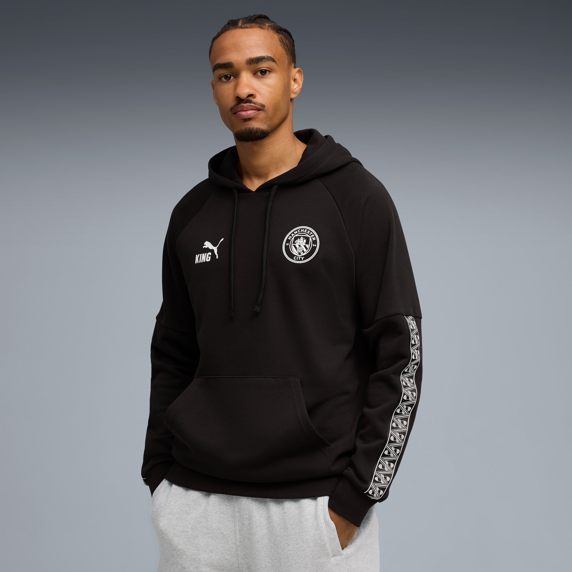 Ein Model posiert vor grauem Hintergrund in einem schwarzen Hoodie mit Teamlogos und grauer Jogginghose.