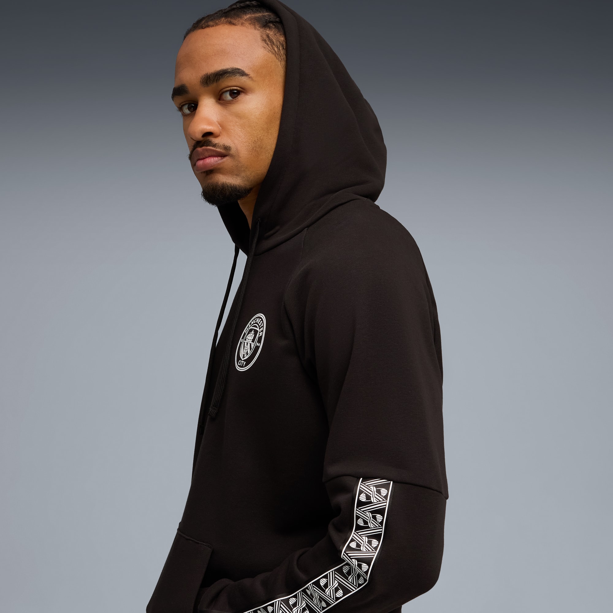 Vor grauem Hintergrund trägt ein Model einen schwarzen Hoodie mit Ärmeldetails und Brustlogo.