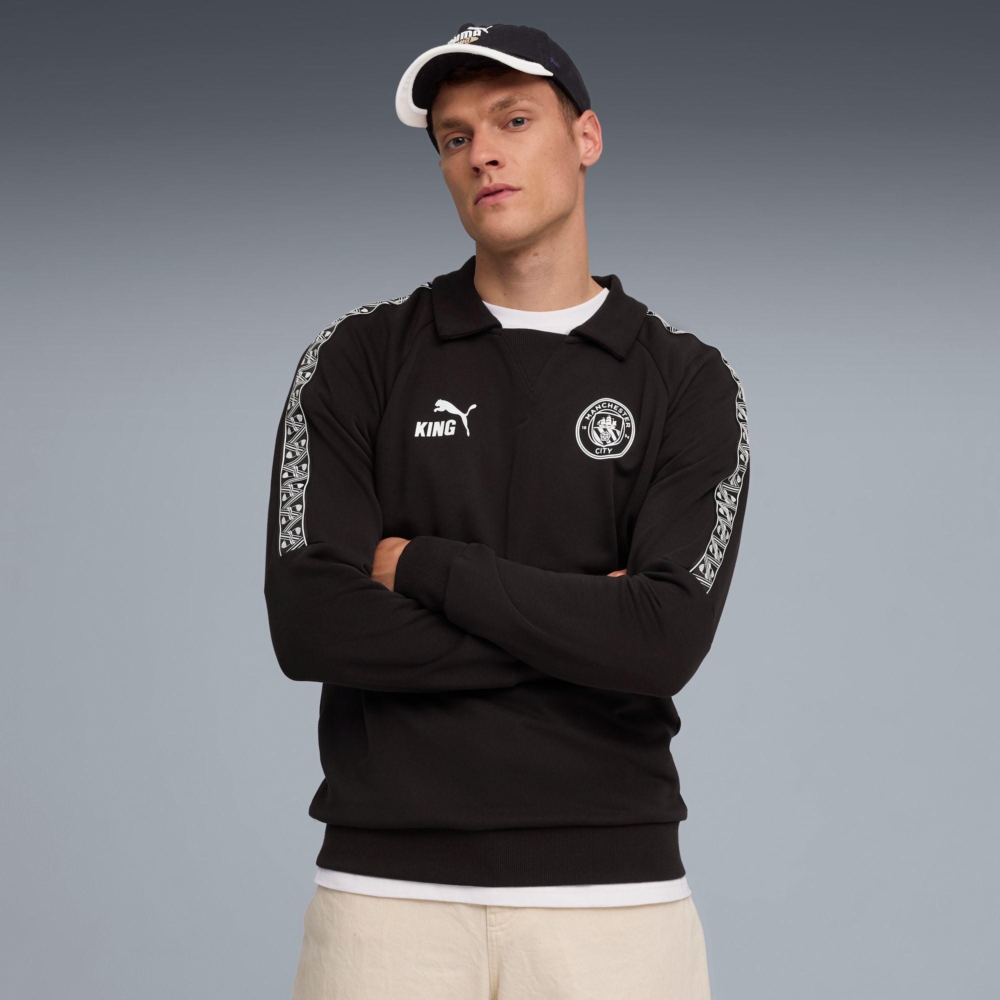 Un modèle, bras croisés, portant un sweat et une casquette noirs sur fond gris.