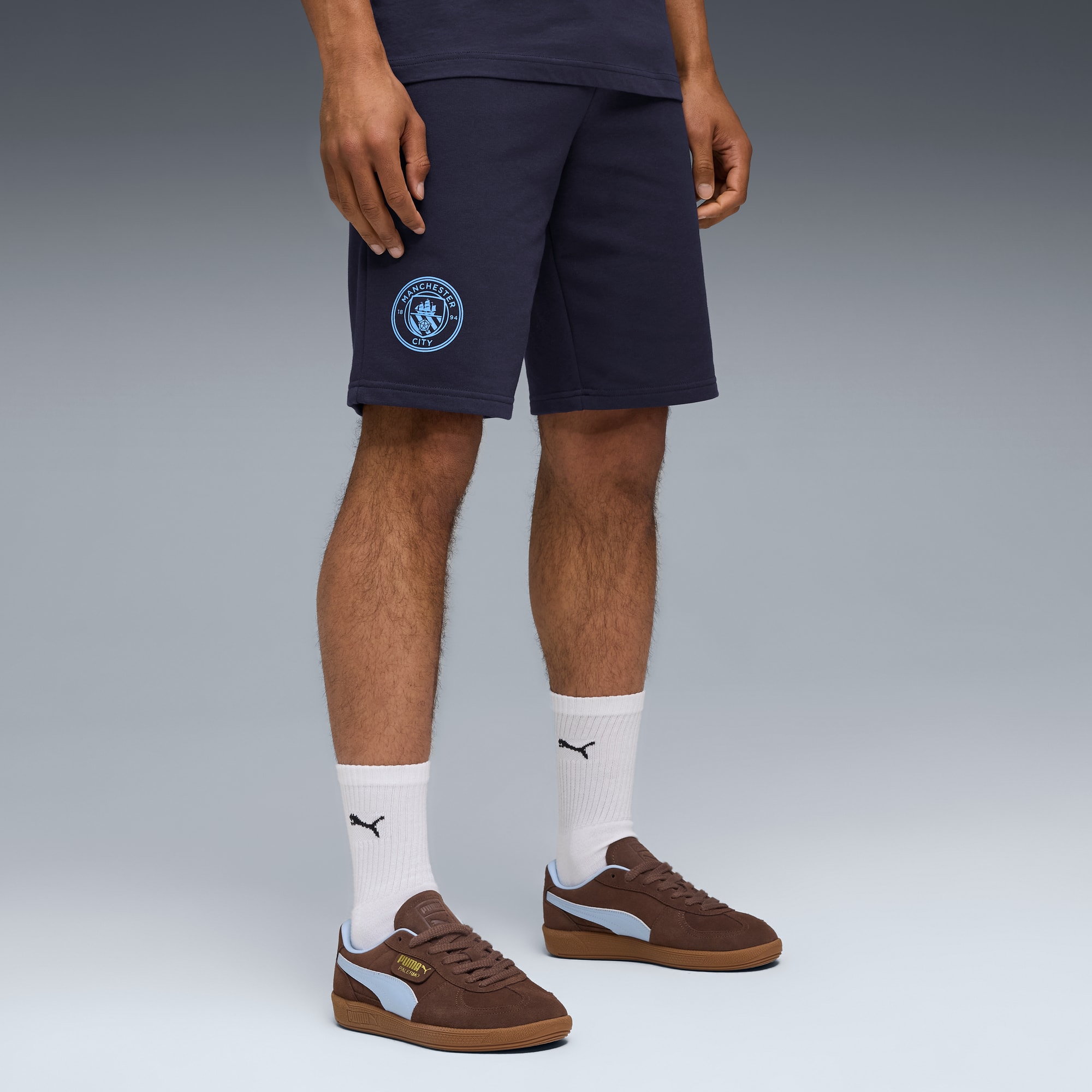 Su sfondo grigio, un modello indossa pantaloncini blu navy, calzini bianchi e scarpe da ginnastica marroni.