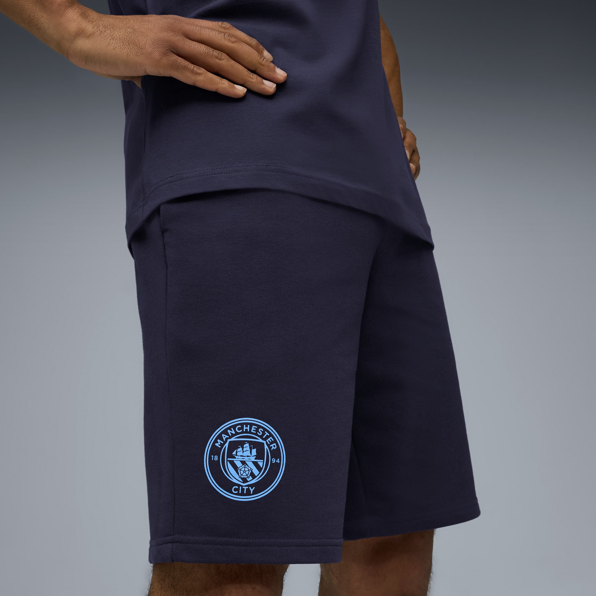 Vor grauem Hintergrund trägt ein Model ein marineblaues T-Shirt und passende Shorts mit Teamlogo.