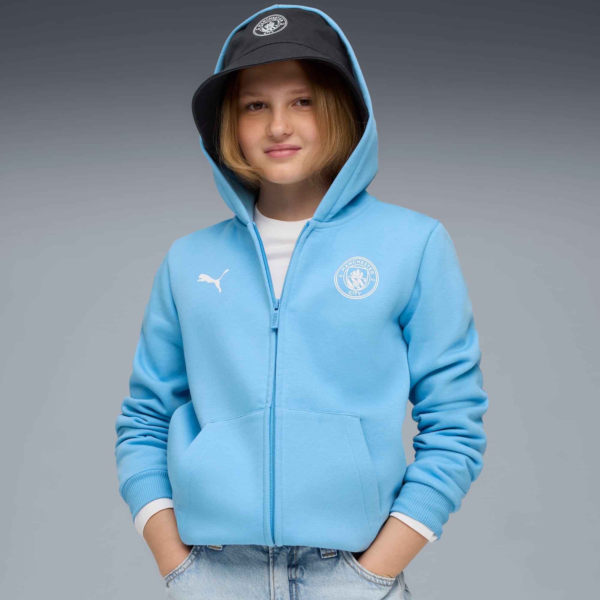 Su sfondo grigio, modello con felpa azzurra con cappuccio, cappello e jeans, mani in tasca.