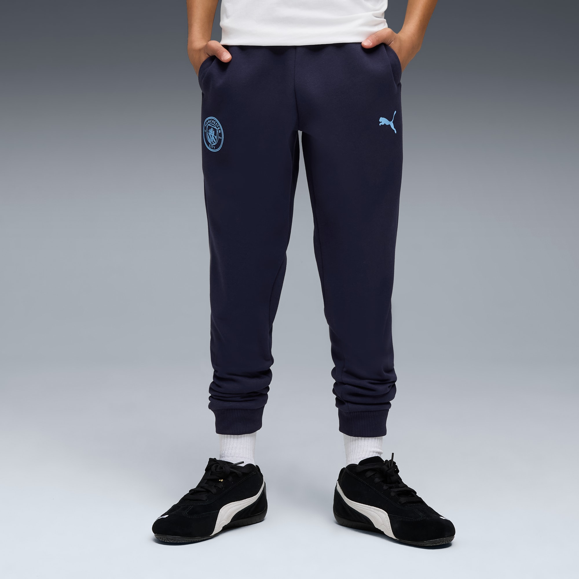 Against grey, a model wears navy joggers with team and brand logos, plus black trainers.