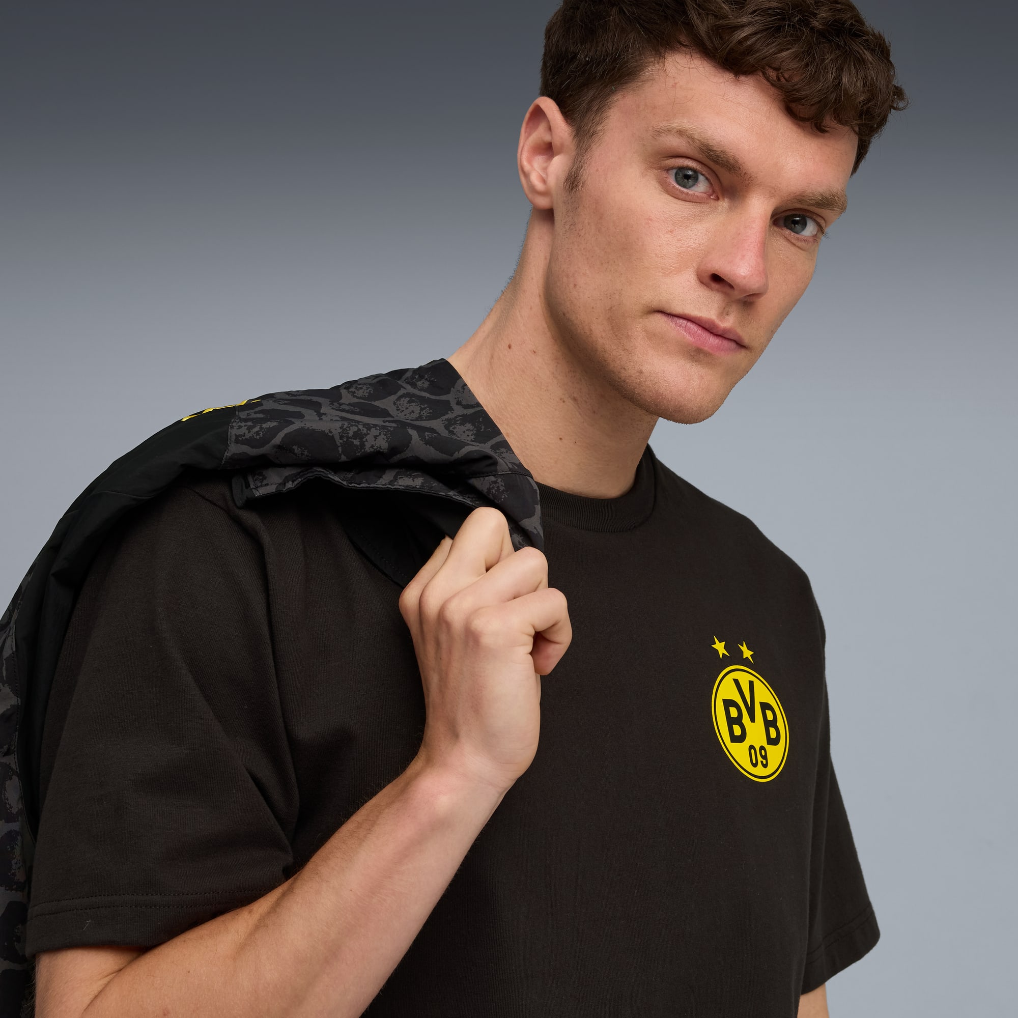 Un modello posa su sfondo grigio, con una t-shirt scura e una giacca sulla spalla.