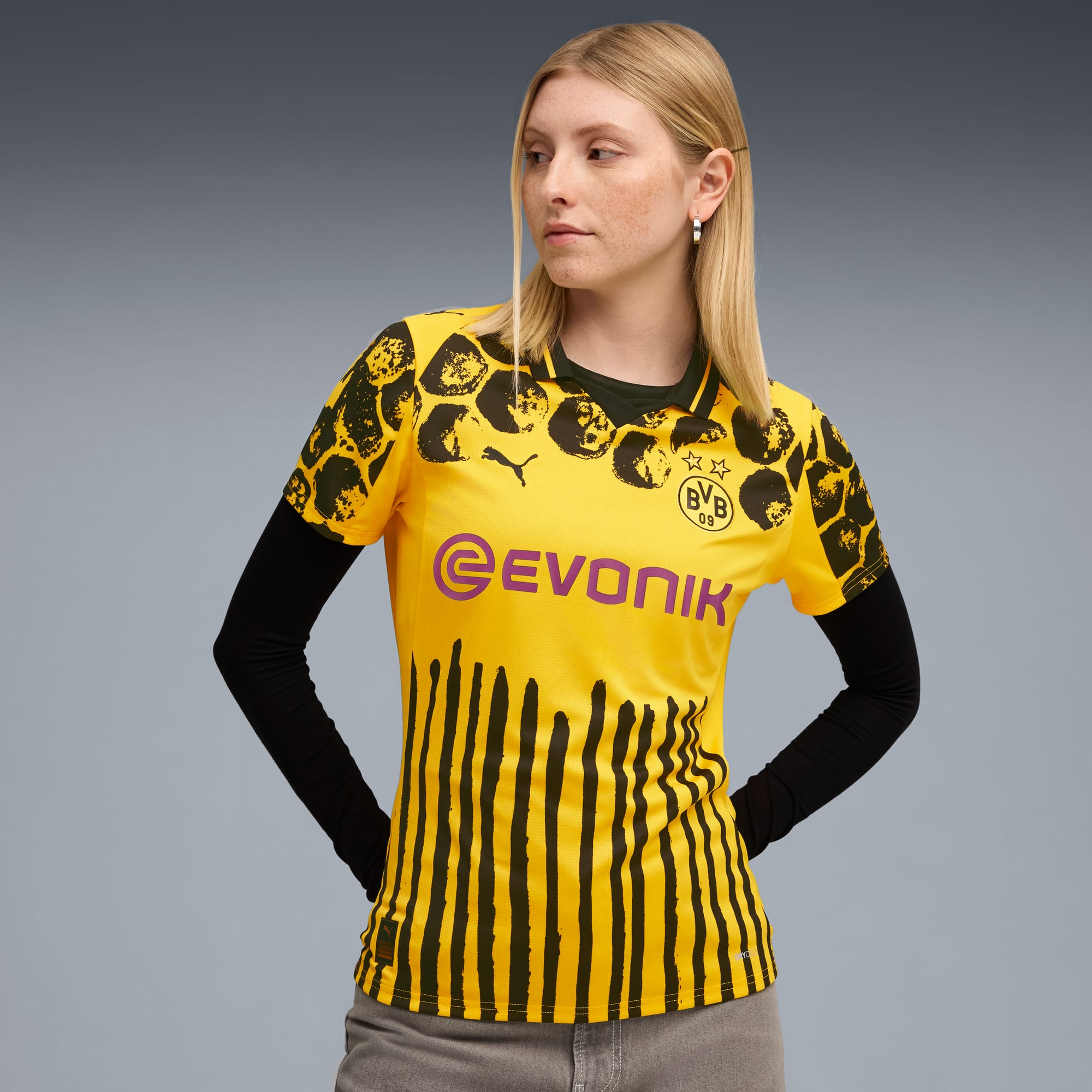 Vor grauem Hintergrund posiert ein Model in einem gelben BVB-Trikot mit schwarzen Ärmeln und grauer Hose.
