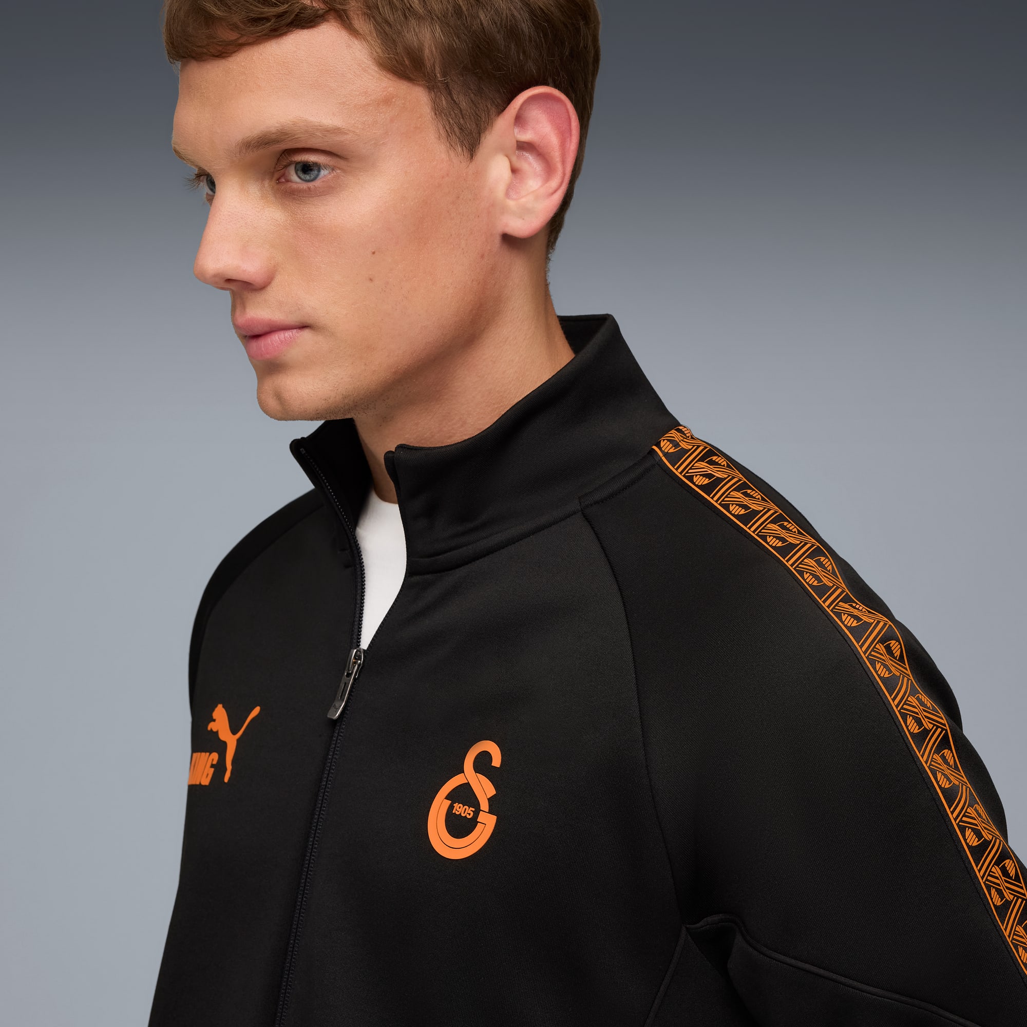 Tegen een grijze achtergrond draagt een model een zwart jasje met oranje accenten en kijkt opzij.