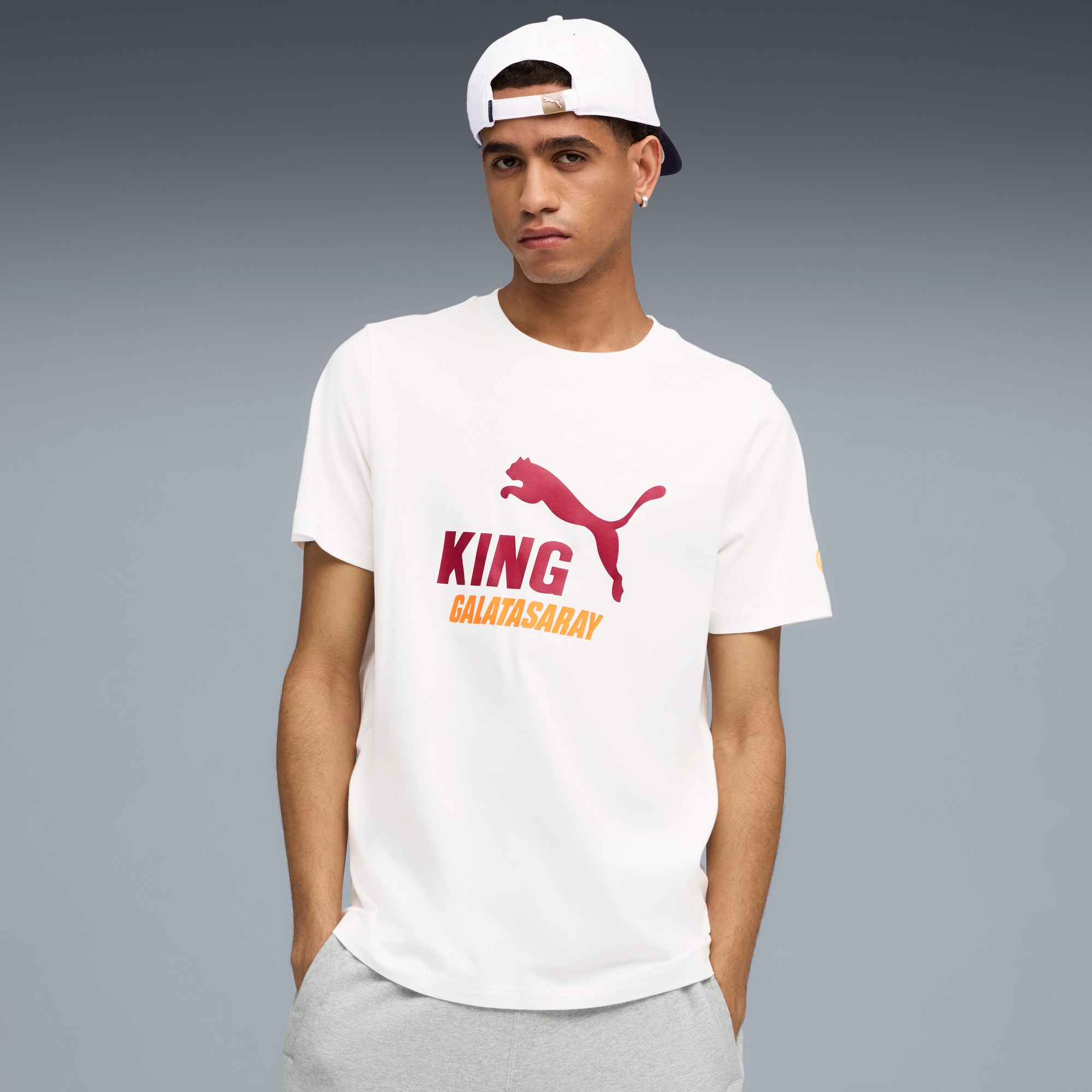 Een model staat tegen een grijze achtergrond, gekleed in een wit t-shirt, grijze korte broek en witte pet.