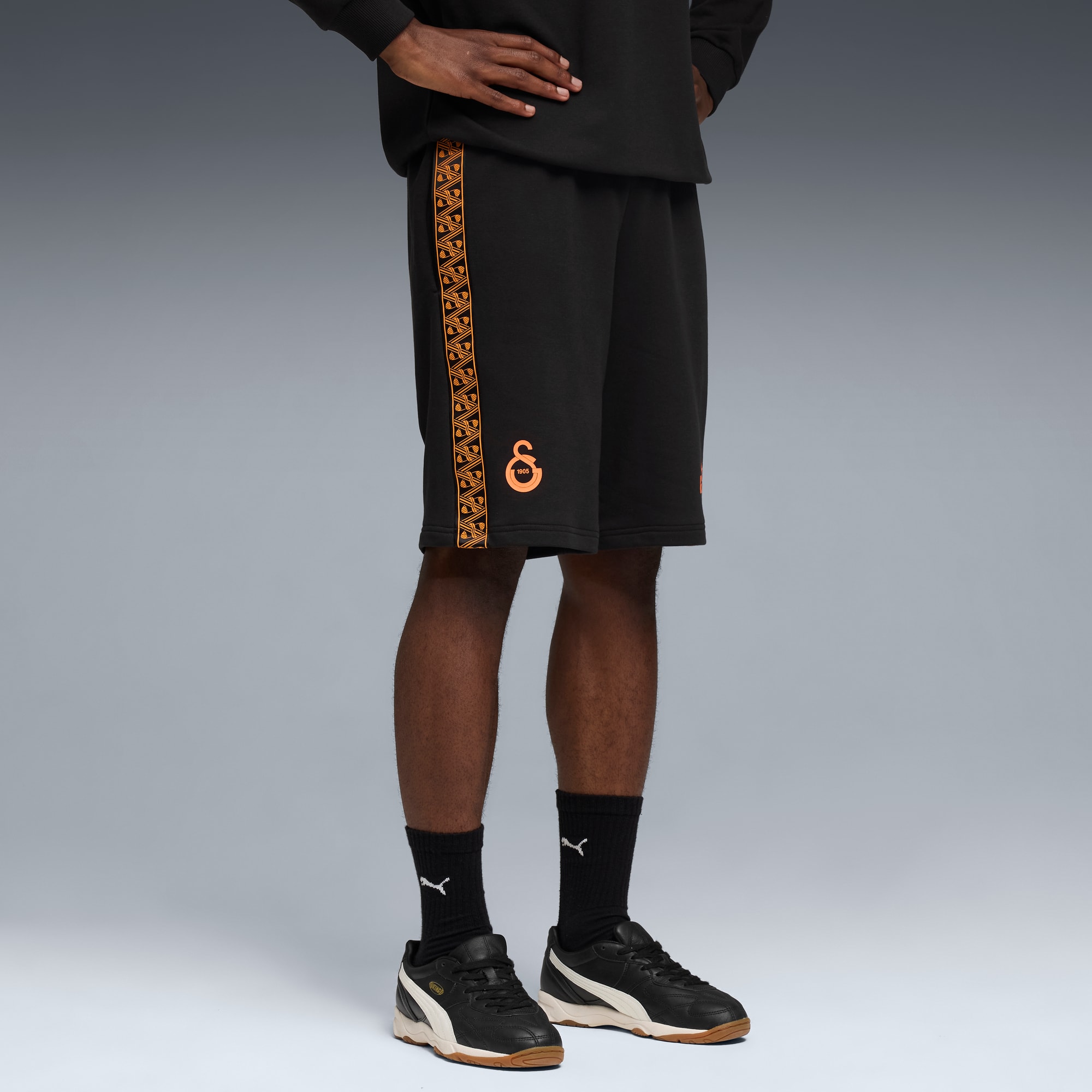 Ein Model posiert vor grauem Hintergrund, trägt schwarze Shorts mit orangen Details und schwarze Turnschuhe.