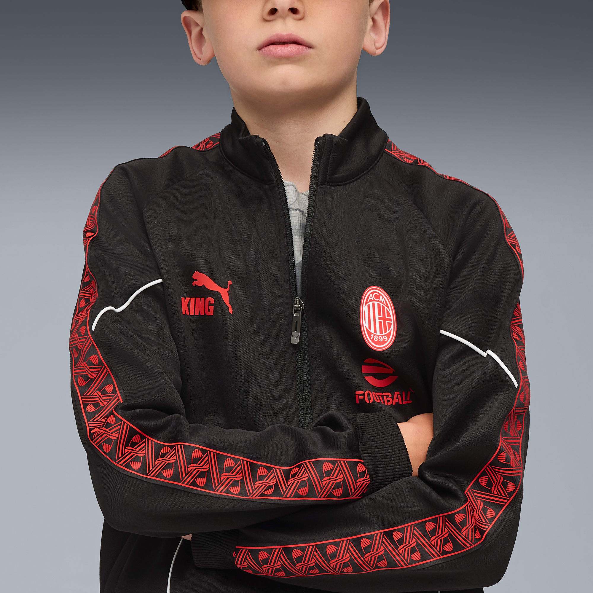 Vor grauem Hintergrund präsentiert ein Kind eine schwarze Jacke mit roten Akzenten und Logos.