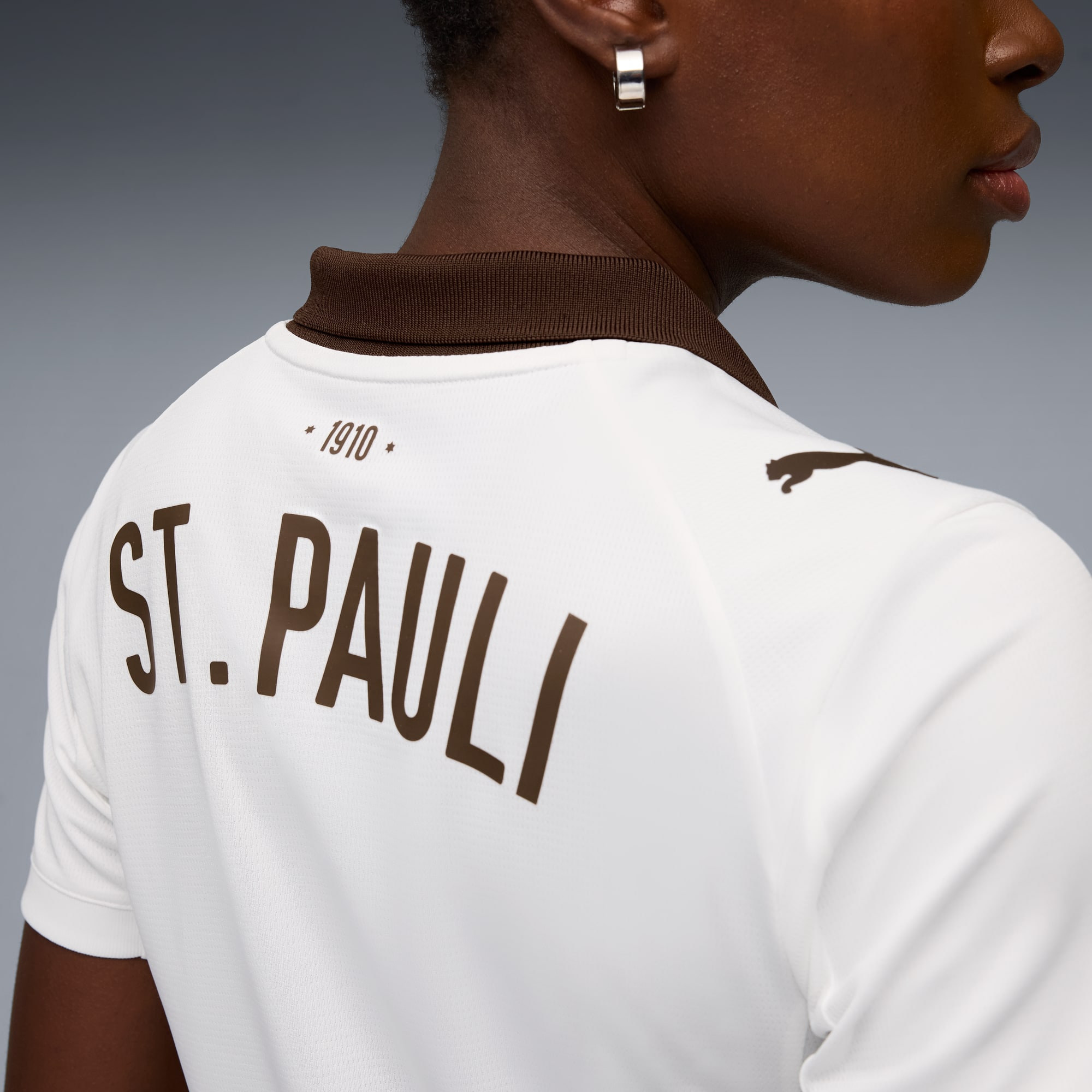 Ein Model trägt ein weißes St. Pauli Trikot mit braunen Akzenten vor grauem Hintergrund.