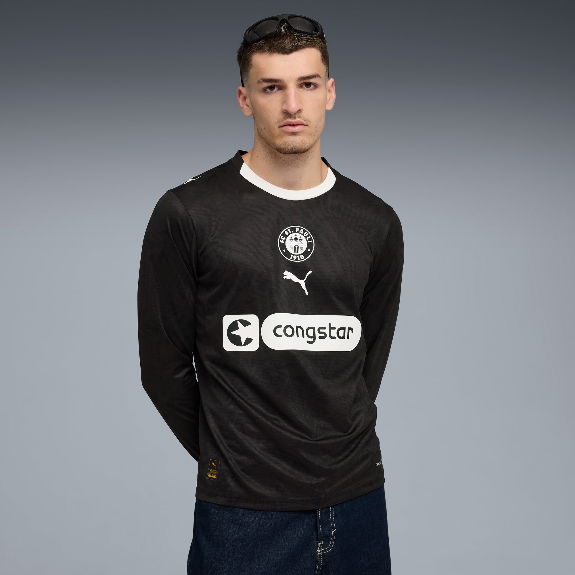 Ein Model posiert vor grauem Hintergrund mit schwarzem St. Pauli-Langarmtrikot und Jeans.