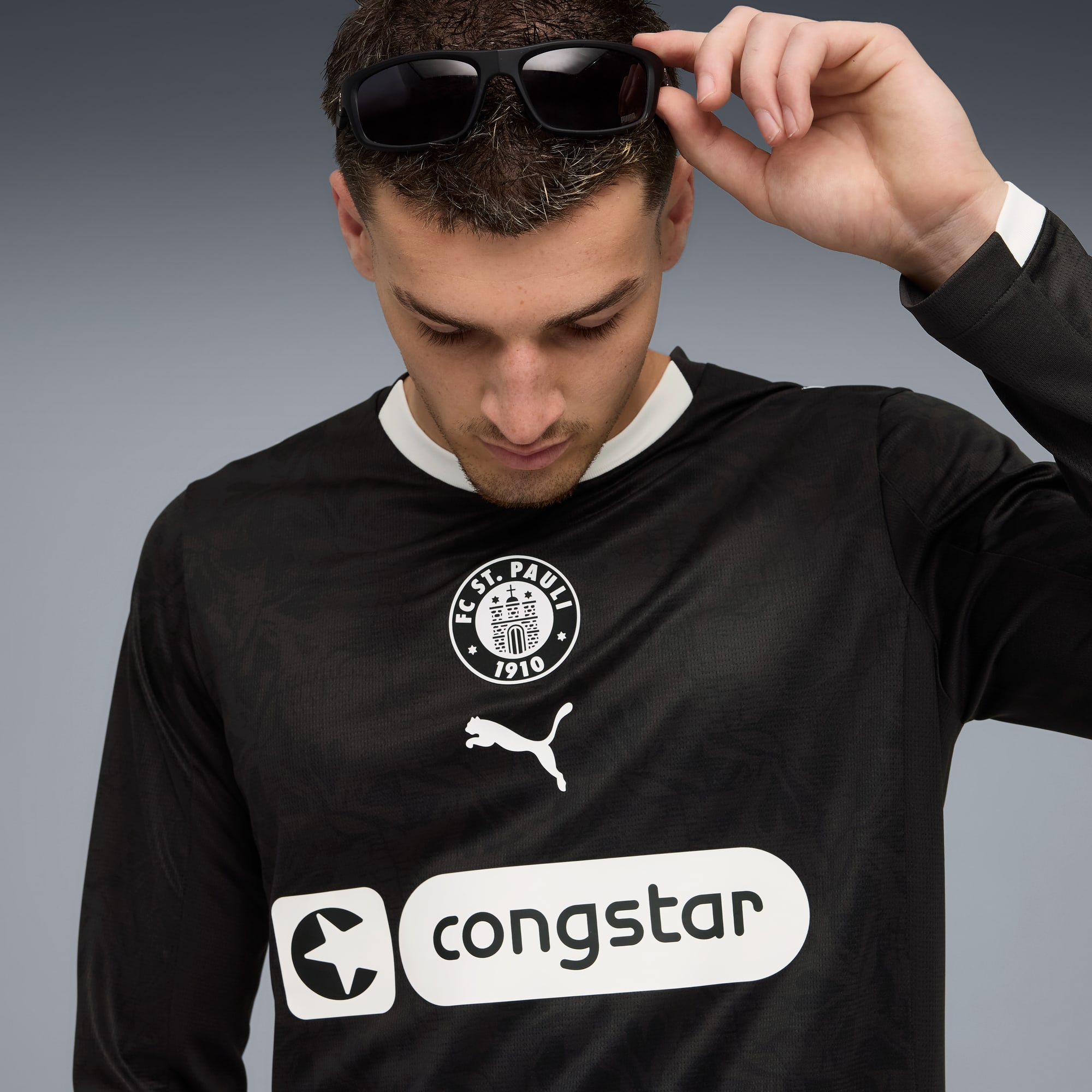 Ein Model justiert eine dunkle Brille vor einem grauen Hintergrund und trägt ein schwarzes F.C. St. Pauli Langarmshirt.