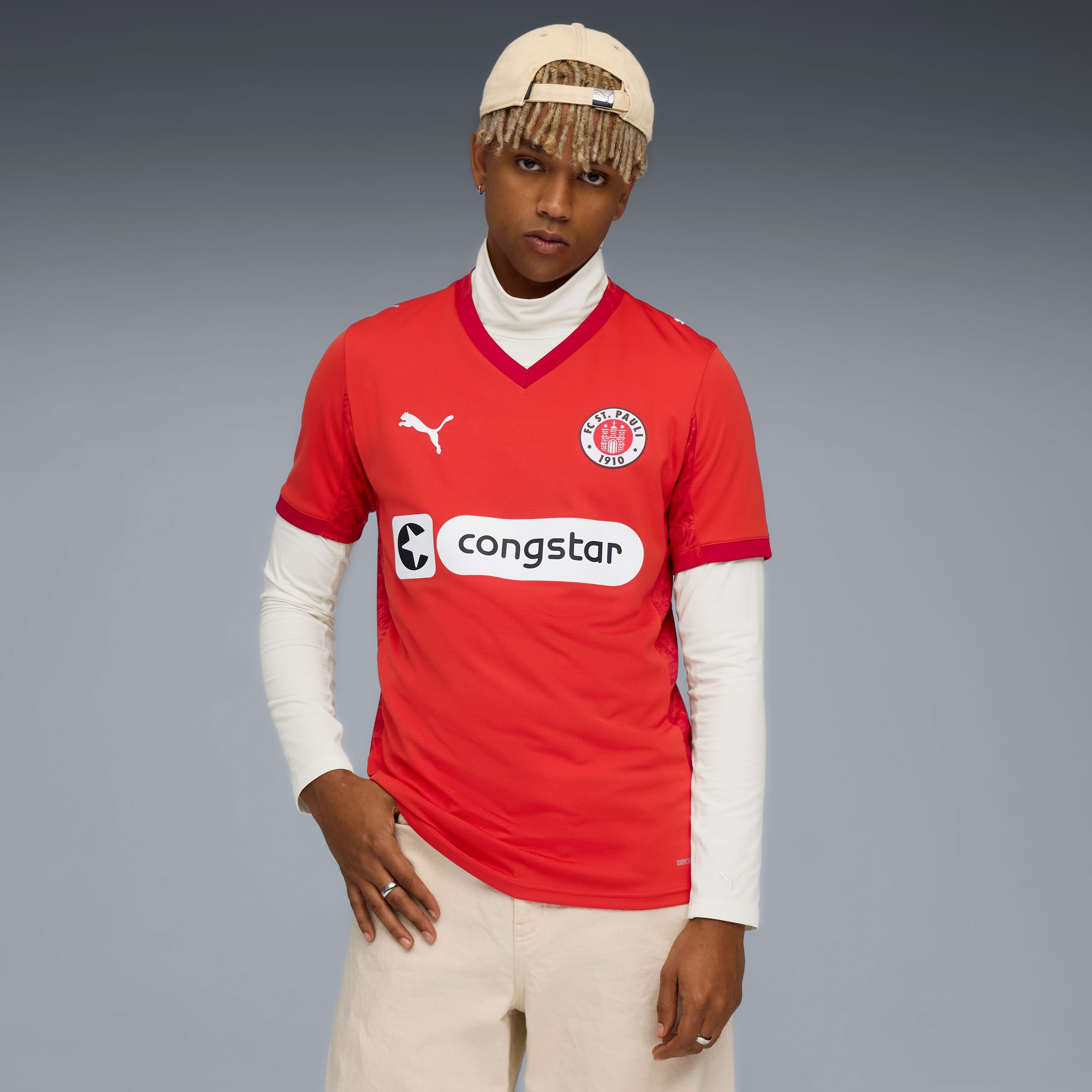 Vor grauem Hintergrund posiert ein Model in rotem FC St. Pauli-Shirt, Hose und Kappe.