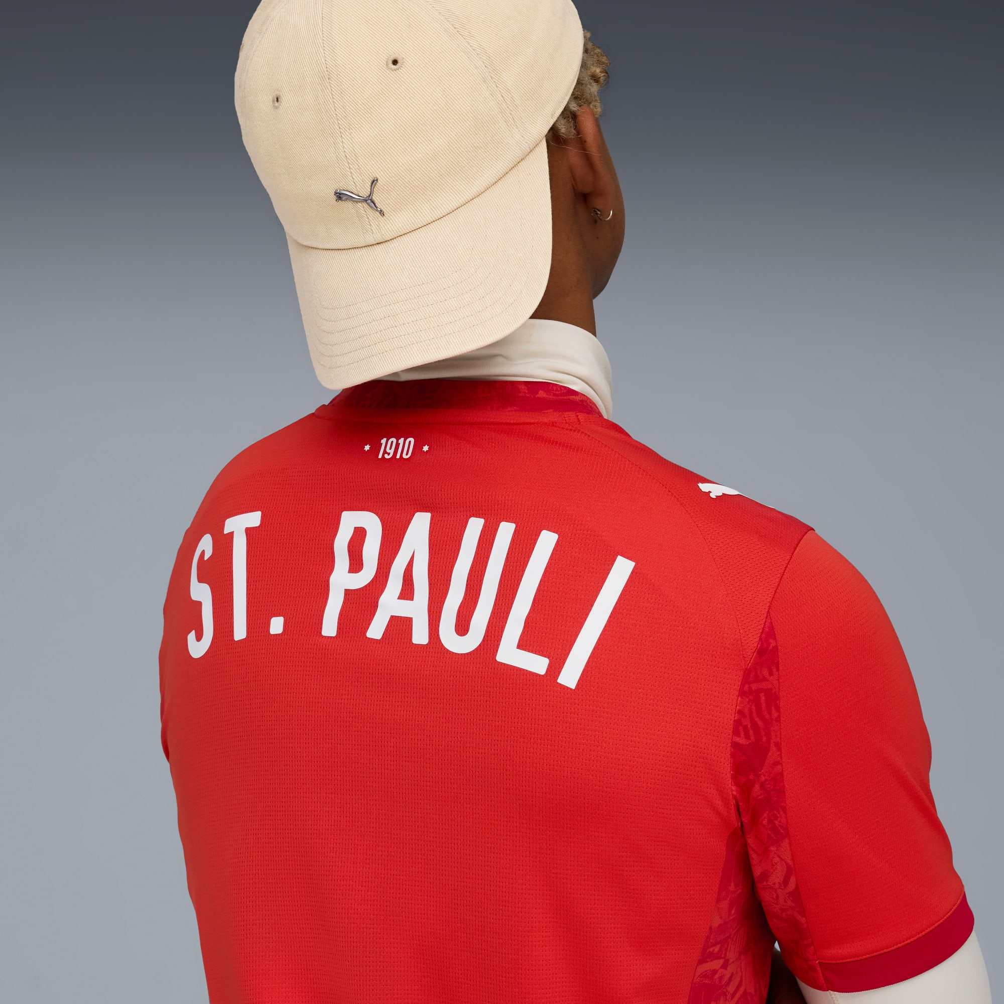Ein Model mit Kappe und rotem St. Pauli-Trikot vor grauem Hintergrund.
