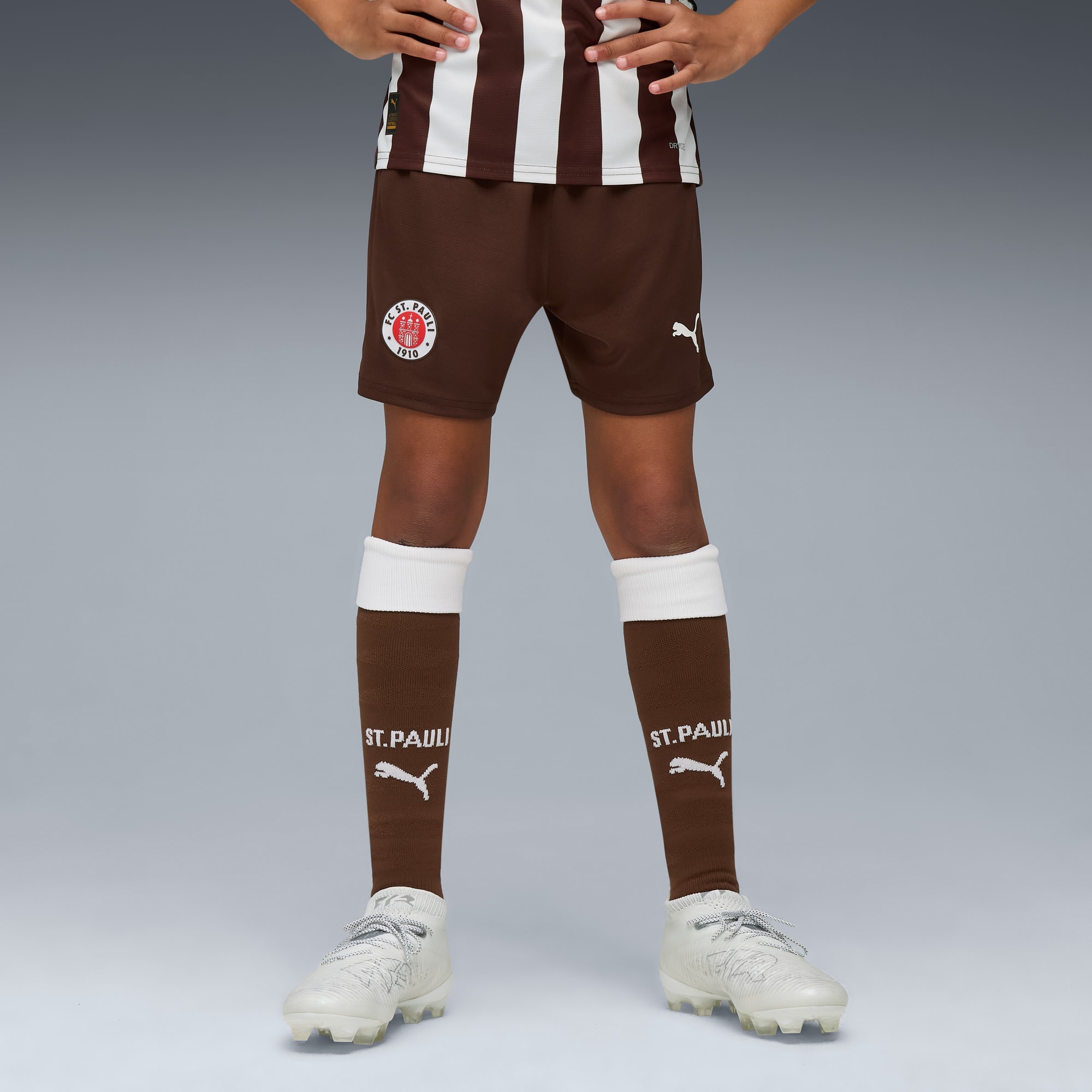 Op grijs draagt een model een St. Pauli-tenue: bruine shorts, sokken en witte hardloopschoenen.