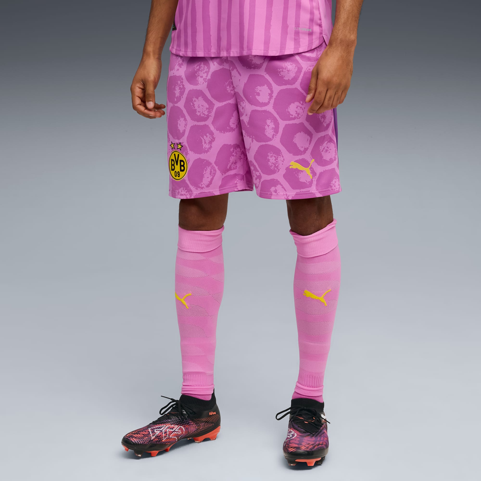 Vor grauem Hintergrund trägt ein Model pinke BVB-Fussballbekleidung mit passenden Socken und Fussballschuhen.