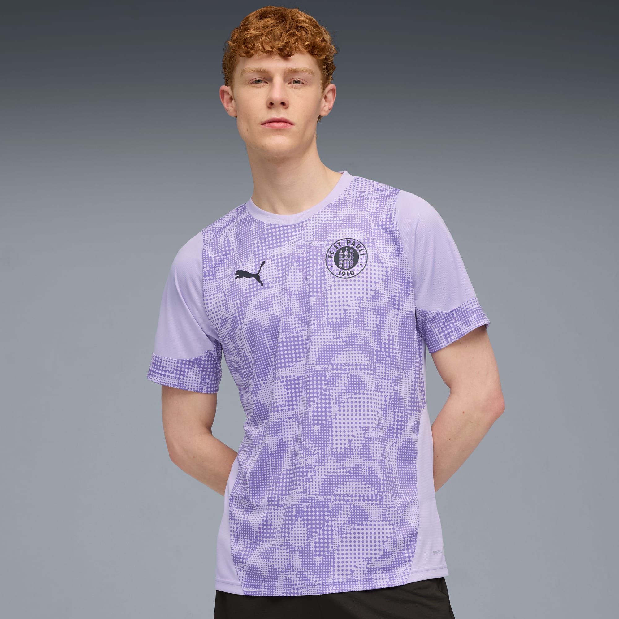 Ein Model steht vor grauem Hintergrund und trägt ein lila gemustertes T-Shirt und Shorts.
