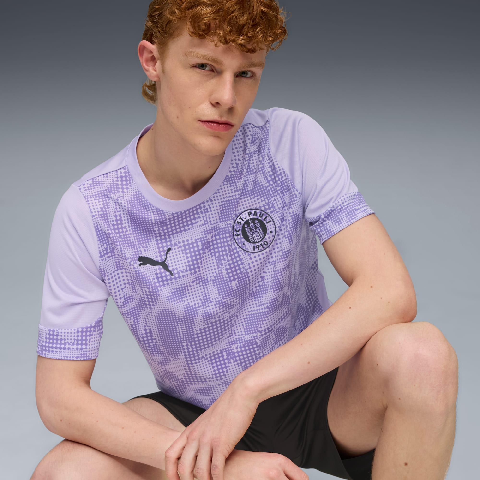 Ein Model posiert vor grauem Hintergrund mit lila Muster-T-Shirt und schwarzen Shorts.