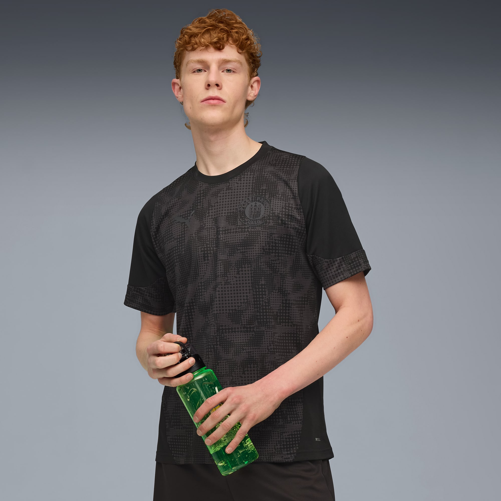 A model against grey holds a green bottle, wearing a patterned black t-shirt and shorts.