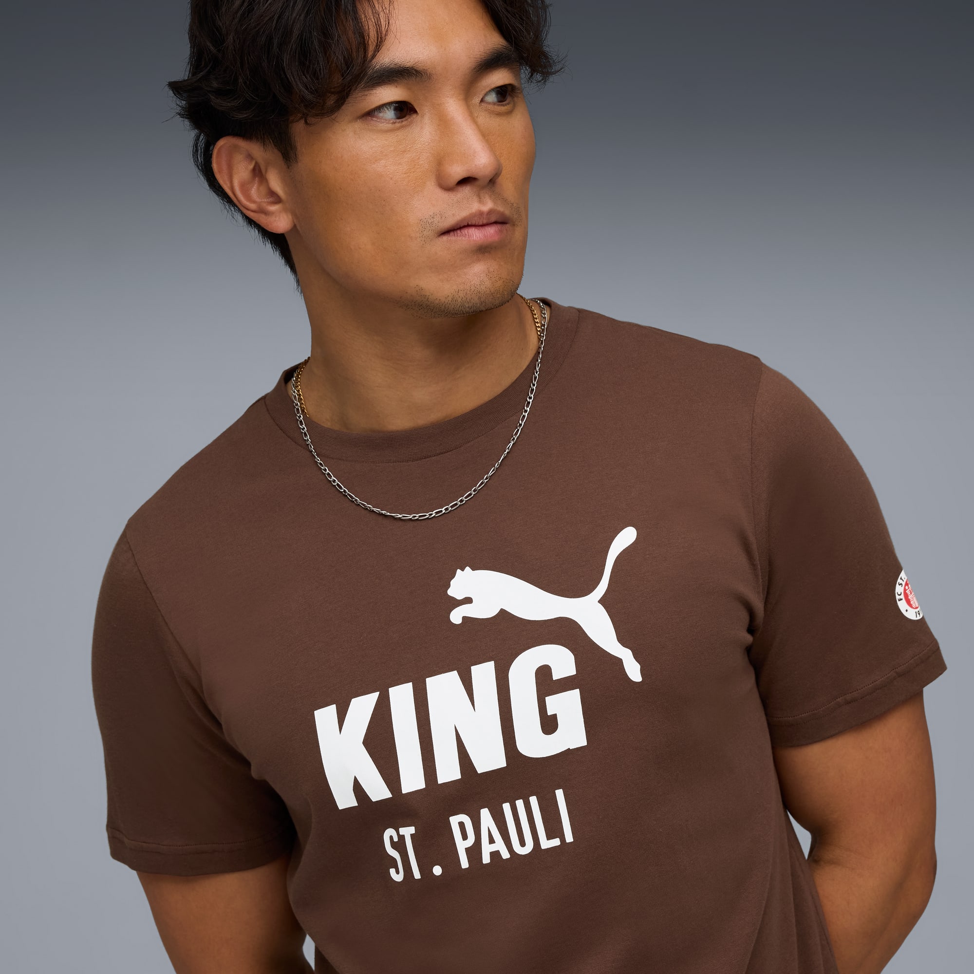 Un modello con catena posa su sfondo grigio indossando una t-shirt marrone con grafica.