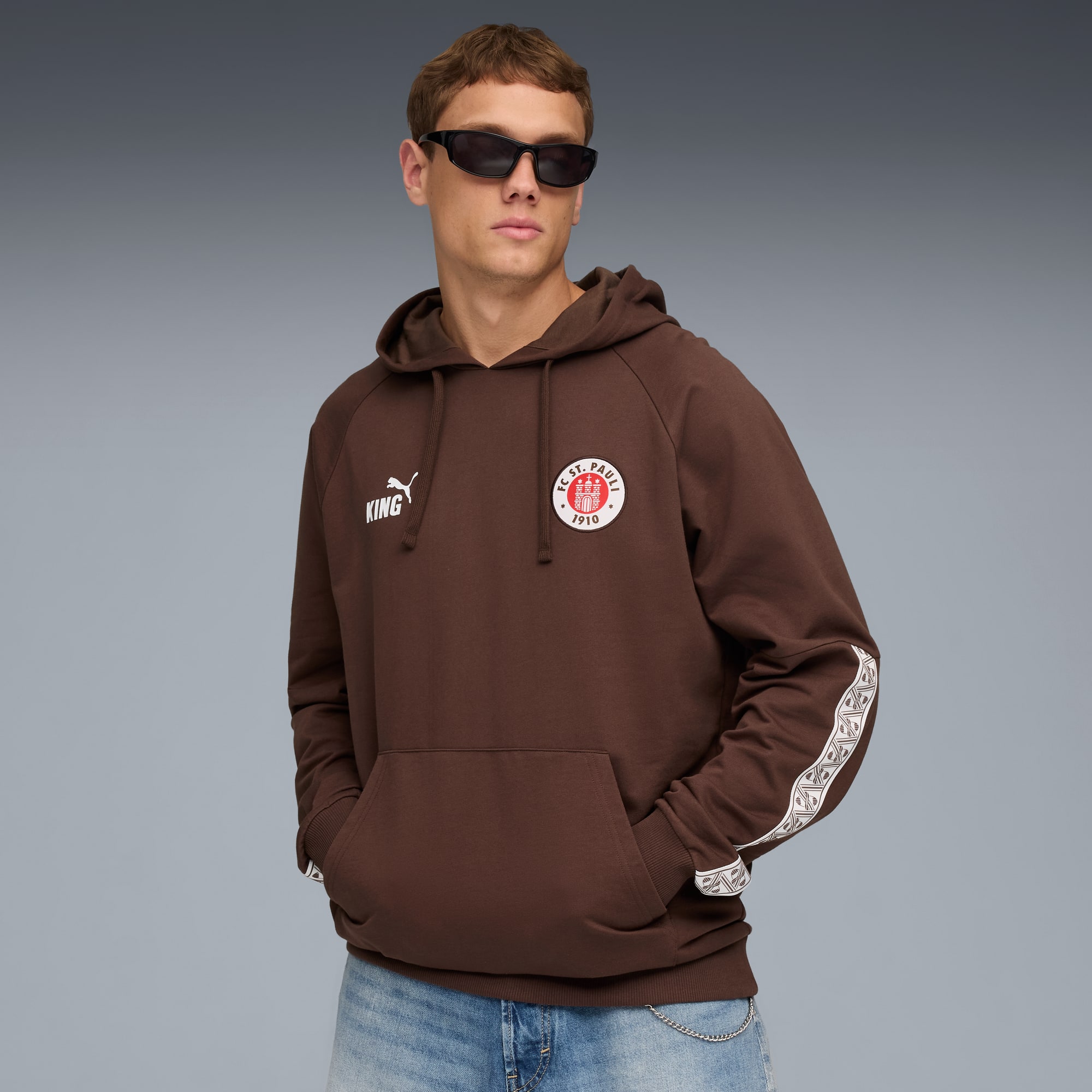 Een model poseert tegen een grijze achtergrond, met zonnebril en bruine hoodie.