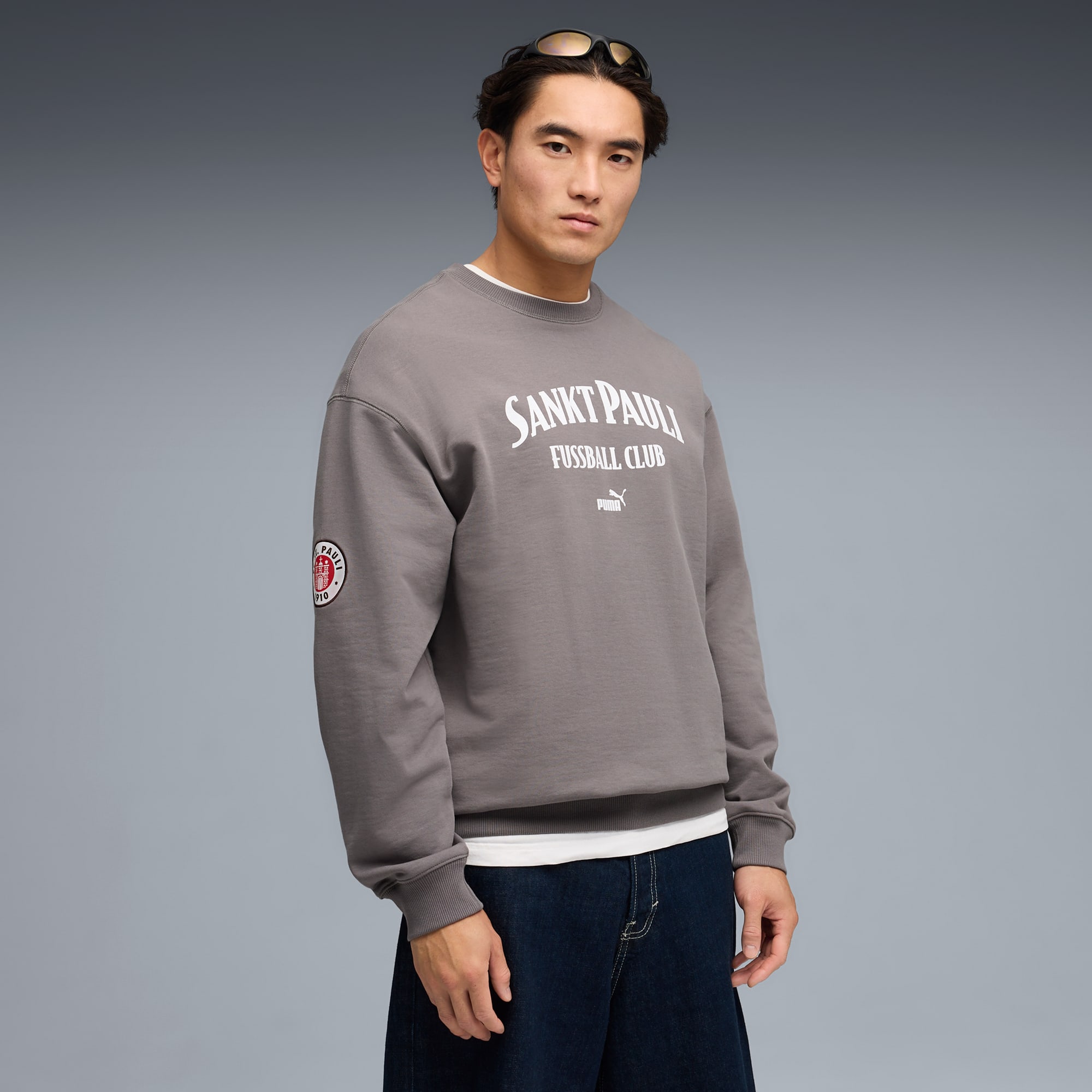 A model poses against a grey backdrop, wearing a grey Sankt Pauli sweatshirt and dark jeans.