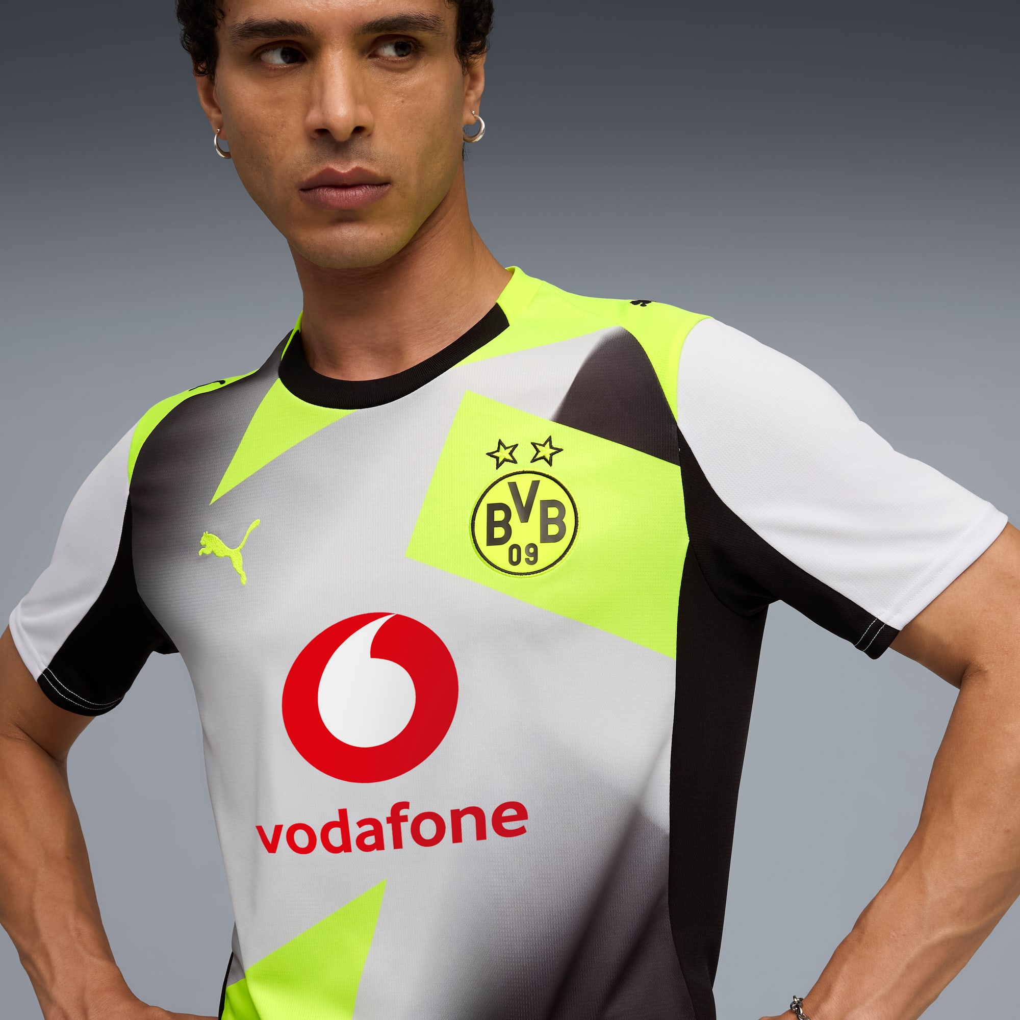 Su sfondo grigio, un modello posa con una maglia del BVB con loghi di sponsor e dettagli verde neon.