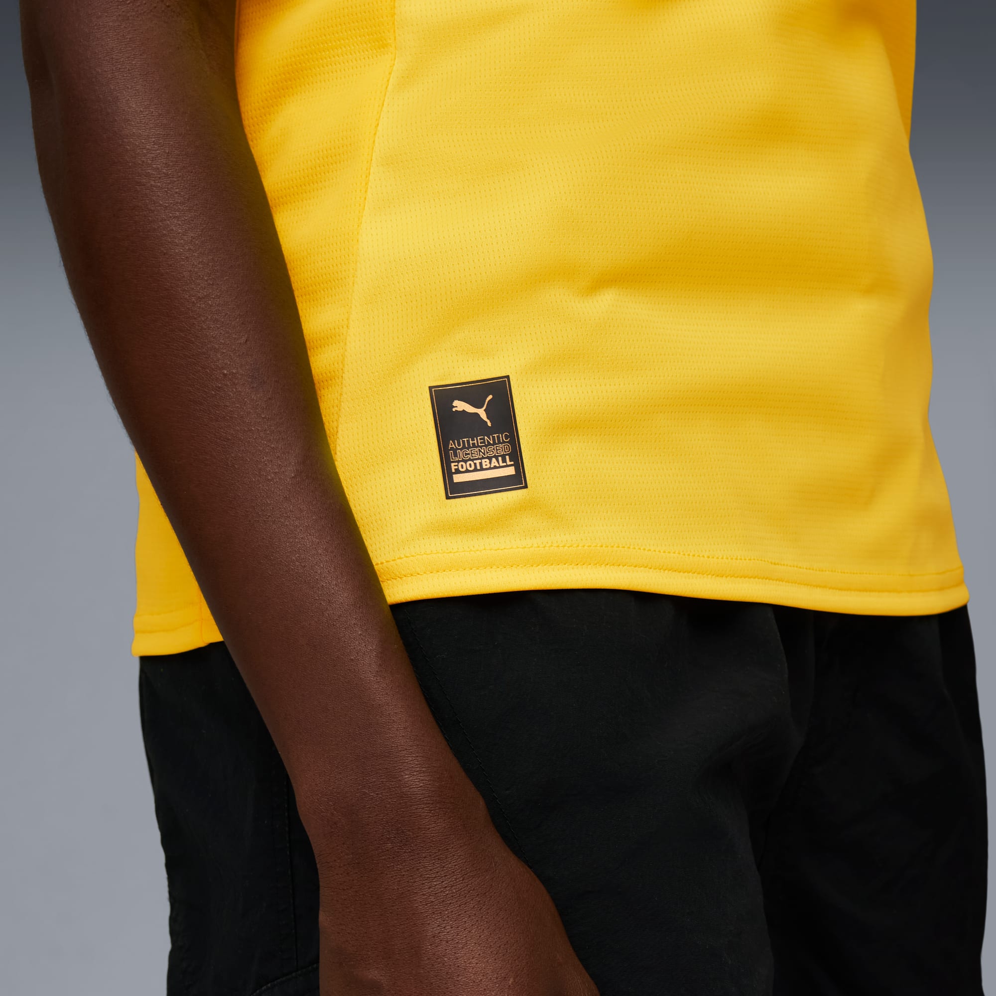 Een model draagt een geel T-shirt met een Puma-logo en zwarte shorts tegen een grijze achtergrond.