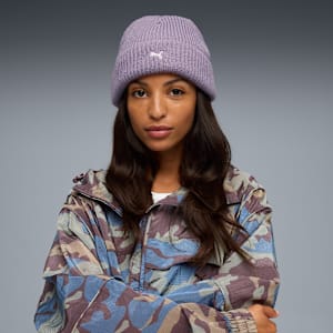 Against grey, a model poses in a lilac hat and camouflage jacket, arms crossed.