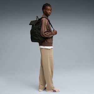Against grey, a model poses with a dark rucksack, brown jacket, beige trousers, and trainers.