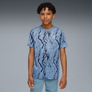 A model with curly hair stands against a grey backdrop, wearing a patterned blue t-shirt and blue bottoms.