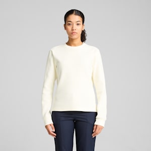 Against a grey background, a model poses in a textured cream jumper and navy trousers.