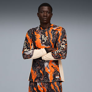 A model with crossed arms, wearing an orange and black patterned long-sleeved shirt, stands against grey.