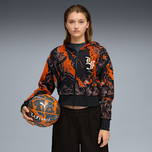 Model with a basketball against grey backdrop, wearing patterned zip hoodie and dark trousers.