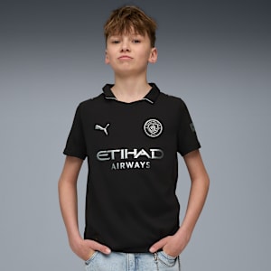 On grey, a model poses with hands in pockets, wearing a black Manchester City football shirt.