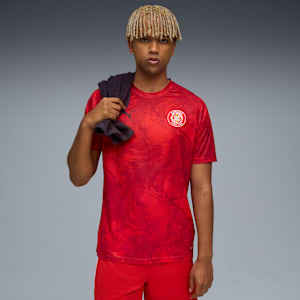 A model poses against grey, wearing a red shirt and shorts, with a towel draped over his shoulder.