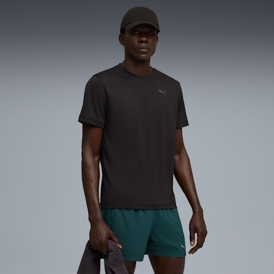 Against grey, a model poses wearing a black t-shirt, cap and green shorts, holding a towel.