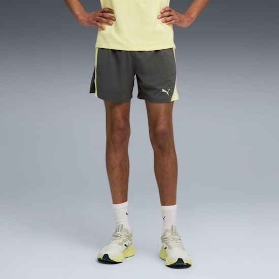 On grey, a model poses in a yellow top, grey shorts with a logo, white socks and running shoes.