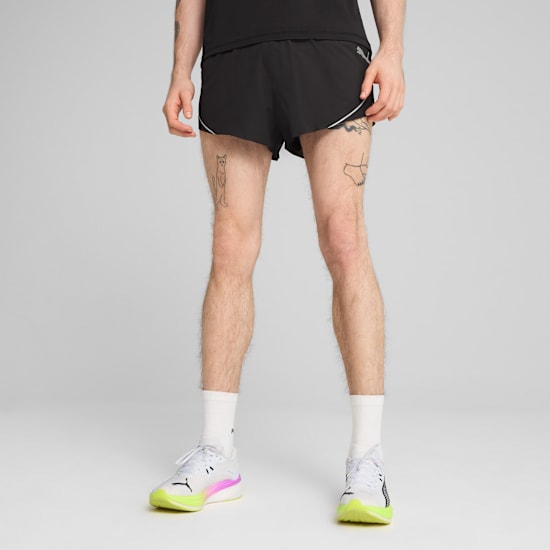 A model in front of a grey background wearing black shorts and running shoes.