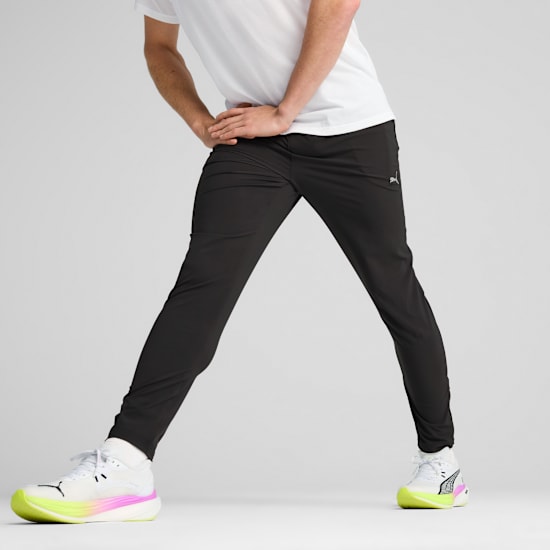 A model stretches on a grey backdrop, wearing a white t-shirt, black trousers, and running shoes.