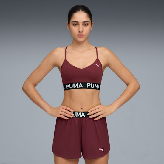 Against grey, a model poses with hands on hips, wearing a maroon bra and shorts.