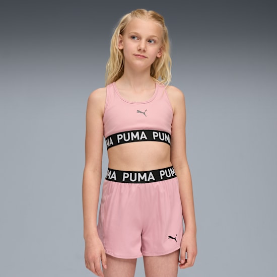 A model is posing against a grey backdrop, wearing a pink Puma crop top and shorts.