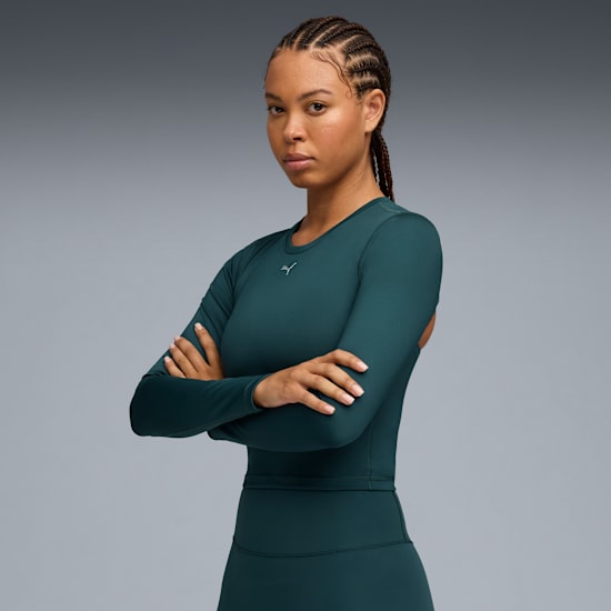 A model with braided hair poses with arms crossed against a grey backdrop, wearing a green long-sleeved top.